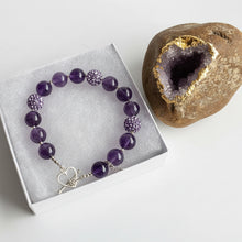 Load image into Gallery viewer, Amethyst  and purple rhinestone bracelet with a silver heart clasp on a white box, next to a decorative rock with a geode.
