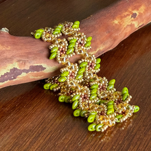 Load image into Gallery viewer, A handmade beaded bracelet with green seed beads and gold-toned metal beads, displayed on a curved piece of driftwood over a dark wooden surface.
