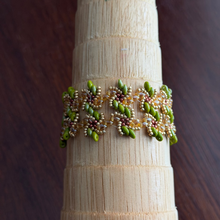 Load image into Gallery viewer, A beaded bracelet with green, gold, and copper seed beads arranged in a vortex pattern, displayed on a light wood cone stand against a dark wood background.
